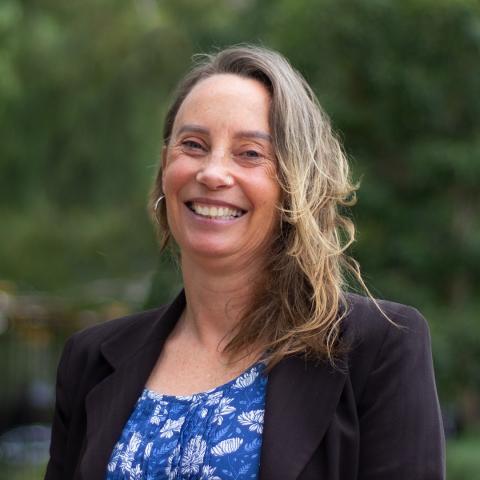 This is an image of Rochelle, a warmly smiling woman in an outdoor environment. Rochelle has light-brown, almost blonde hair that falls below her shoulders and is tucked so you can see one of her hoop earrings. She wears a dark blazer, blue floral blouse and a nose ring.