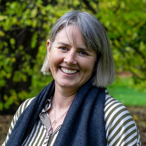 This is an image of Izzy who warmly smiles at the camera. She's in a white and oliver green striped blouse with a charcoal scarf around her neck and she stands in a bright green park environment. She has salt and pepper hair cut just above the chin with a side-swept fringe.